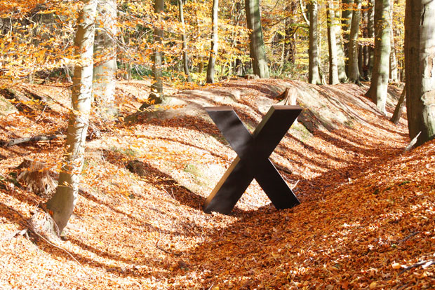 Ein großes, massives, schwarzes X, fast mannshoch, steht in einem sonnigen Herbstlaubwald neben einem Bodenwall.