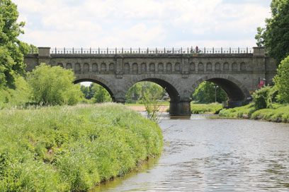 Bräunlicher Fluss mit grasbewachsener grüner Böschung mit einzelnen Büschen. Über den Fluss wölbt sich eine graue steinerne Brücke mit 3 Bogen und einem schwarzen metallenen Handlauf auf den Seitenwänden.