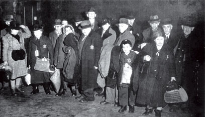 18 Personen, darunter ein Kind, eng beieinanderstehend mit Taschen und Beuteln in den Händen, mehrheitlich mit Hüten auf dem Kopf. Auf der linken Brust eingenähte „Judensterne“.