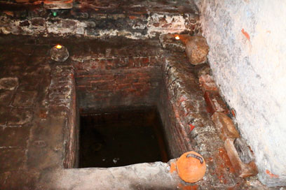 Rechteckiger Einlass aus Klinkern im Boden, der mit wenig Wasser gefüllt ist. An der linken und rechten oberen Ecke Teelichter, an der rechten unteren Ecke ein Gewicht.