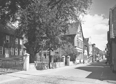 Schwarz-weiß Fotografie einer Straße mit Kopfsteinpflaster. Die Häuser links und rechts sind zweistöckig. Das linke Haus steht etwas zurückgesetzt; davor ein großer Baum.