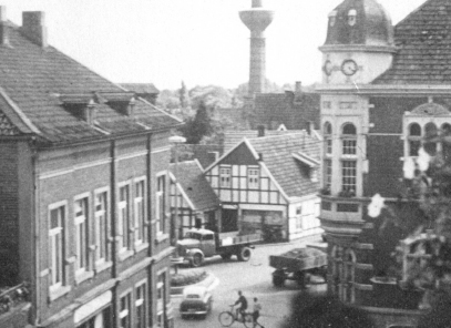 Schwarz-weiß-Fotografie eines Platzes mit Autos und Fahrrädern. Um den Platz verschiedene Gebäude, manche mit Klinker, Fachwerk oder Stuck, im Hintergrund ein Turm.