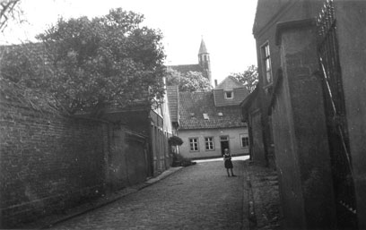 Gepflasterte Straße, links eine Mauer, rechts Mauer und Gebäude, am Ende der Straße ein Kind. Dahinter einstöckiges Gebäude mit Kirchturm darüber.
