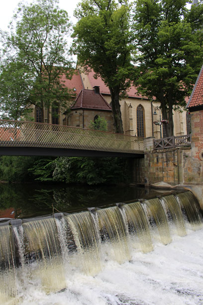 Ein Stauwehr mit fließendem Wasser, unterhalb einer Brücke. Im Hintergrund eine Kirche.