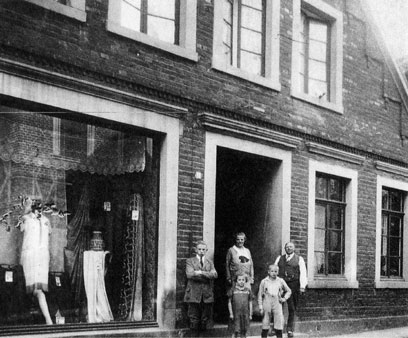 Fünf Personen vor einem großen Eingang eines Gebäudes. Links davon ein Schaufenster mit Schaufensterpuppen und Kleidungsstücken.