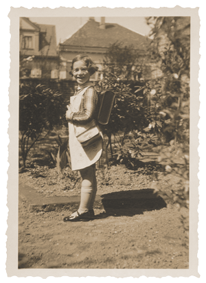 Helga Becker-Leeser vor einer Hecke in Kleid mit kleiner Tasche und Schulranzen.