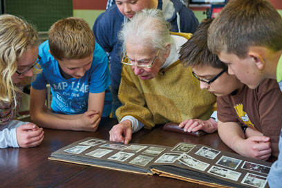 Helga Leeser blättert zusammen mit einigen konzentrierten Kindern in einem Fotoalbum an einem braunen Tisch.