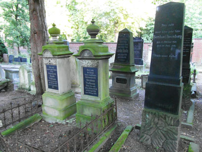 Links zwei umzäunte Grabsteine mit hellem aber bemoostem Stein und dunklen Tafeln, in der Umgebung dunkle Säulen auf hellem Stein.
