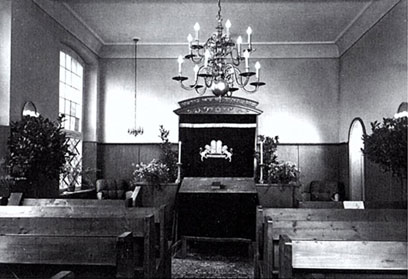 Schwarz-weiß-Fotografie mit Bänken links und rechts, über dem Mittelgang ein kleiner Kronleuchter, vorne ein Toraschrein.