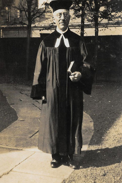 Black-and-white photograph of Hermann Manuel in robe with glasses, holding a book in his left hand. He is standing on a path with large stone slabs.