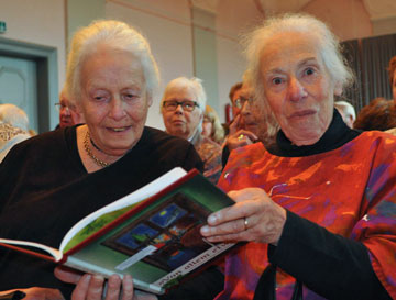 Helga Leeser holds an open book and looks into the camera; Ingrid Leeser looks at the book. Behind them, some people are also sitting.
