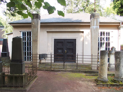 View of one-storey building; directly in front are two high columns and a metal fence; on the right and left, gravestones.